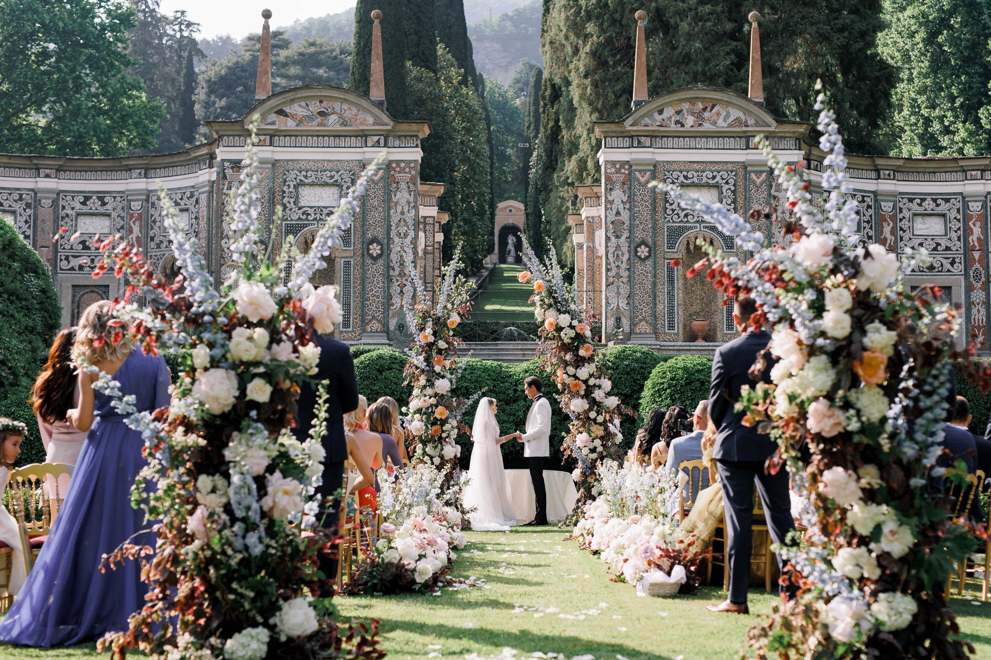 An outdoor wedding ceremony framed by flowers and historic gardens, shown in its entirety